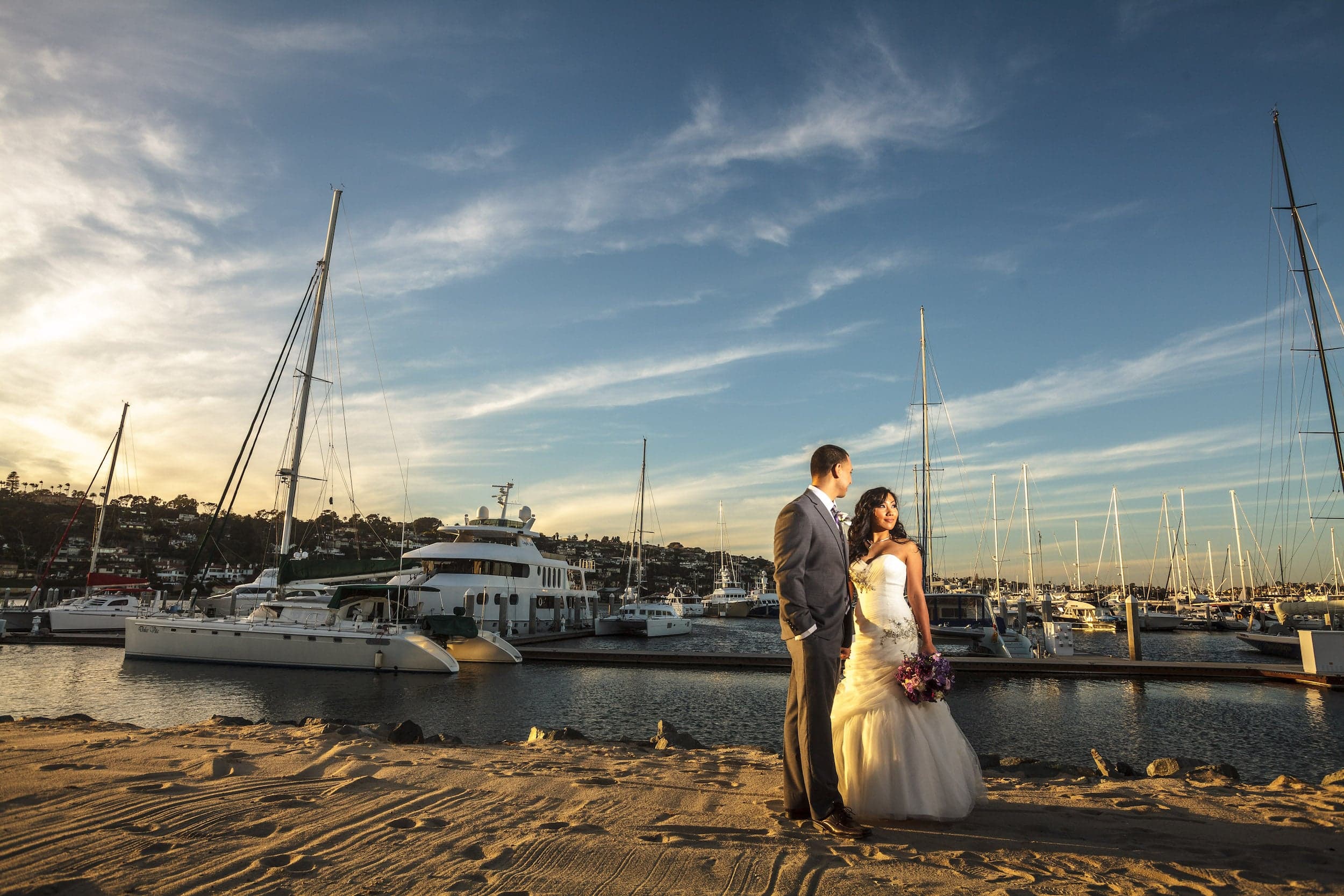 A just married couple embracing for a photo on the beach with boats in the background