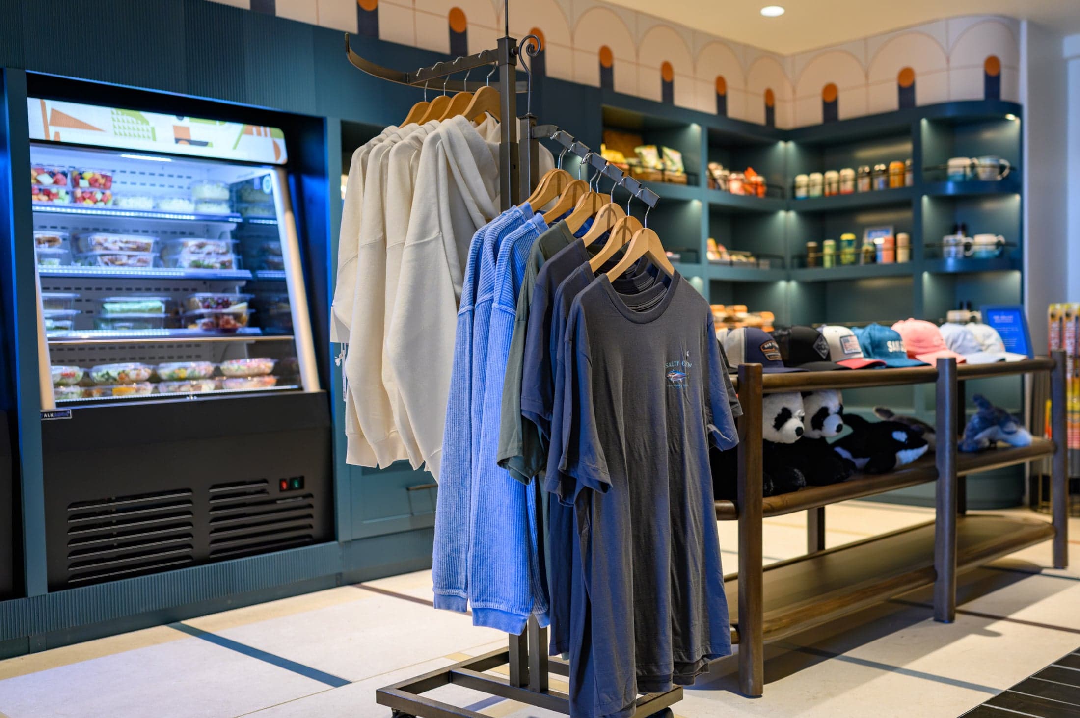 Grab-and-go retail display featuring apparel and refrigerated grab-and-go food case, with hats, plush toys, and snacks on shelving in the background.